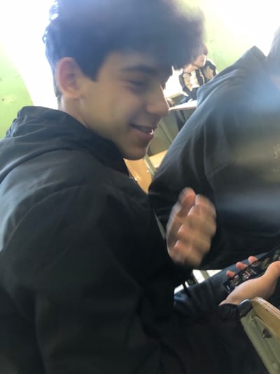 The image depicts an indoor scene in what appears to be a classroom or educational facility, located in Ortabatumi, Georgia.
In the left foreground, a young male individual is captured in a side profile, facing right. He has dark, wavy hair and is wearing a dark-colored, possibly black, hooded jacket. His facial expression indicates a slight smile or smirk, with his mouth partially open. His right hand is blurred, suggesting motion, and is positioned near the mid-section of his body.
To the right, another individual is partially visible, primarily their torso and lap. This person is wearing a dark, possibly grey, hooded garment. They are holding a black smartphone or similar electronic device in their left hand, with their right hand also positioned near the device, appearing to interact with its screen. The screen displays a colorful interface with multiple circular icons.
In the blurred background, a third individual is partially visible, seated at a desk. This person has dark, curly hair and is wearing a horizontally striped top with alternating light and dark bands.
The environment includes a light green wall to the left of the main subject and wooden desks or chairs visible throughout the background. The lighting is bright, with some glare present, particularly in the upper right portion of the image.