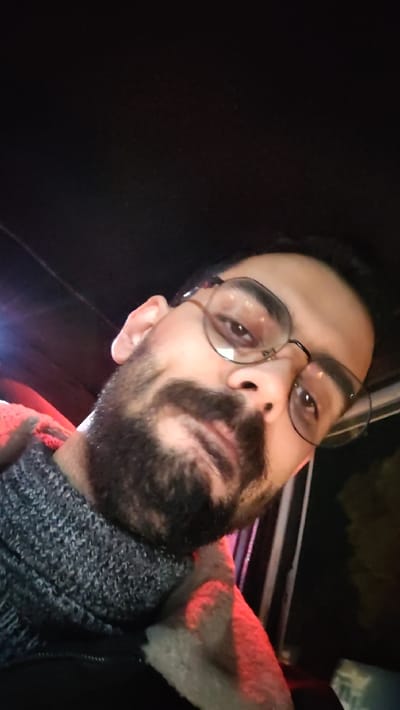 A person with a beard and glasses looks directly at the camera. He's wearing a gray knit sweater with a light-colored collar or lining visible. The background is predominantly dark, with some indistinct red or orange light visible. The lighting suggests an indoor or nighttime setting.