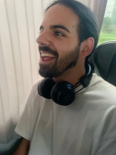 A male individual is depicted in a close-up, chest-up shot, seated inside a vehicle. He has dark, long hair pulled back and a full dark beard. His head is turned towards the right side of the frame, and his mouth is open in a wide smile, revealing teeth, consistent with laughter. He wears a plain white short-sleeved t-shirt. Black over-ear headphones are suspended around his neck. The background on the left features vertical white or light-colored window blinds. To the right, blurred green foliage is visible through a window, and a dark grey vehicle seat headrest is partially visible. The specific location within the city of Leiria, Portugal, is not discernible from the visual evidence presented in the image, other than the subject being within a transport vehicle.