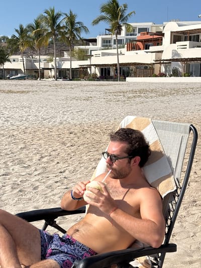 A man with brown hair, a beard, and sunglasses lies on a striped beach chair, drinking from a coconut with a straw. He wears blue and pink patterned swim trunks. A red string bracelet is visible on his left wrist. The chair is positioned on a light-colored sandy beach. In the background, white buildings with varying architectural styles are visible, along with palm trees. The sky is clear and blue. The location appears to be a beach in La Paz, Mexico, based on the architectural style and the presence of palm trees.