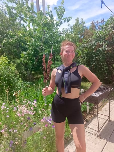 A woman stands in a backyard, holding two skewers of cooked meat. She is wearing a black sports bra and black shorts, with a dark scarf tied around her shoulders. In the background, there is a charcoal grill, lush green trees, and a patch of colorful wildflowers. The sky is blue with scattered clouds, suggesting a sunny day. The overall atmosphere is relaxed and casual, hinting at an outdoor meal or gathering. The woman appears happy and content. The location is identified as Vyazovka, Russia.