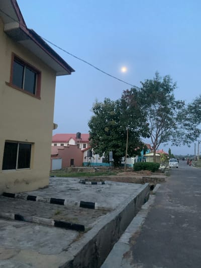The image presents an outdoor street scene in Osu, Nigeria, observed during twilight, indicated by a bright, full moon against a light blue sky. In the immediate foreground to the left, a portion of a two-story building with a light beige facade and brown-trimmed windows is visible. A concrete slab extends from its base, featuring black and white painted curb stops marking a designated area. A concrete drainage ditch runs along the right edge of this slab, parallel to a paved road that recedes into the distance on the right. A thin, dark cable crosses the upper left quadrant of the sky.
In the midground, a large tree with dense green foliage stands prominently. Beyond it, various residential or commercial structures are discernible, including a building with a reddish roof topped by a black water tank, and another building painted white and blue. On the paved road, a white van or minibus is parked or moving slowly on the right side. Further down the road, several indistinct human figures are visible in the distance. The overall environment suggests a suburban or semi-urban setting with infrastructure including roads, buildings, and natural vegetation.