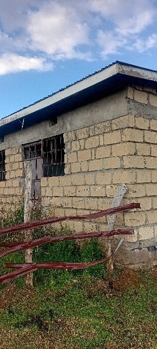 This image depicts the exterior of an unfinished or weathered single-story building in Kinale ward, Kenya. The building features walls constructed from light-colored concrete blocks, with visible mortar lines. Portions of the wall, particularly near the top, appear to have a rough cement finish or an incomplete plaster layer. The roof has a dark, corrugated or tiled surface with a prominent white fascia board. Two rectangular window openings are visible on the building's side, each fitted with dark, possibly metal, security bars that appear discolored or rusted. One window opening also contains what appears to be a partially boarded-up or broken wooden frame.

In front of the building, a rudimentary fence or barrier is present, constructed from several horizontal, rust-colored metal strips attached to two vertical wooden posts. A light-colored concrete slab or broken piece of masonry rests against the building's wall near this barrier. The ground in the foreground is covered with patches of green grass interspersed with areas of dry soil and sparse vegetation. The sky above is light blue with scattered white cumulus clouds. No individuals or active interactions are visible within the scene.