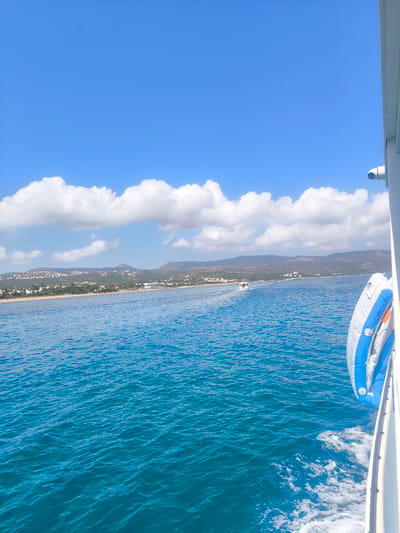 A high-angle, long shot captures a bright, sunny day at sea. The vast expanse of the Mediterranean Sea, rendered in vibrant turquoise, fills the lower half of the frame, its surface textured by gentle waves and disturbed by the wake of a vessel. On the right, a portion of a white boat is visible, its railing and a white and blue life preserver prominently featured. In the distance, a coastal town, characterized by white buildings and lush greenery, stretches along a bay, backed by rolling hills under a brilliant blue sky dotted with fluffy white clouds. A small boat can be seen in the middle distance. The scene is characteristic of the coastline near Polis Chrysochous, Cyprus, a popular tourist destination known for its beautiful beaches and clear waters.