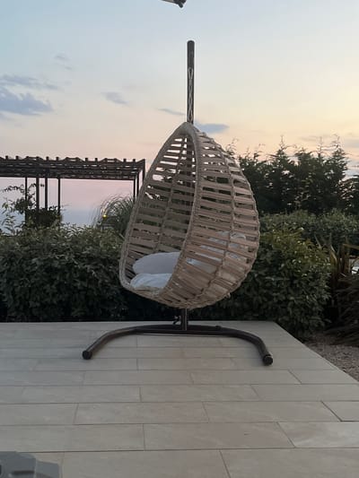A hanging rattan chair with a white cushion sits on a tiled patio. The chair is suspended from a dark metal stand, which curves out to rest on the light-colored tiles. Behind the chair, lush green bushes and trees form a natural backdrop, with a wooden pergola structure visible to the left. The sky above is a soft gradient of pale blue and pink, suggesting either dawn or dusk. The overall scene evokes a sense of relaxation and tranquility in an outdoor setting. The image appears to have been taken in Lastva Grbaljska, Montenegro. No people or text are visible in the frame.