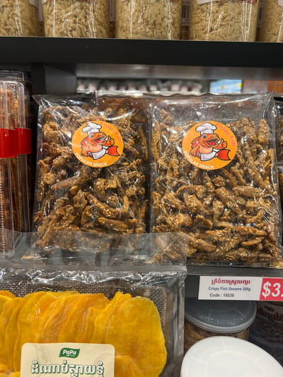 The image shows a retail display shelf in a store, likely a supermarket or convenience store, in Siem Reap, Cambodia. The shelf is stocked with various food products. In the foreground, there are packages of dried mango slices. Behind the mango, there are two clear plastic bags containing what appears to be crispy fish snacks coated in sesame seeds. Each bag has an orange circular sticker with a cartoon fish wearing a chef's hat, and text that reads "Crispy Fish" and "POPULAR SNACK". To the right of the snack bags, a price tag is visible, indicating "Crispy Fish Sesame 200g" and a code "16036", with a price of "$3." and what appears to be Cambodian Riel currency. Further back on the shelf, several clear jars filled with a dry, shredded product are visible, along with other packaged goods. The lighting suggests an indoor environment.