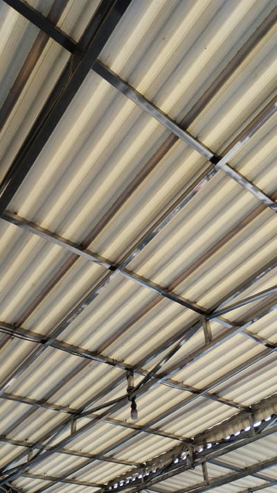 An upward angled shot shows a corrugated translucent roofing material supported by a dark metal frame. The roofing panels have alternating bands of light beige and pale gray, creating a striped pattern. The metal frame consists of intersecting beams and supports that form a grid structure overhead. One light fixture, with a single bulb, hangs from a crossbeam in the center of the frame. The scene appears to be an outdoor covered area, possibly a market or a pavilion, in Tanjungsari, Indonesia. There are no people visible in the image. The lighting is diffused, suggesting an overcast day or a shaded location. The overall impression is one of utilitarian construction.