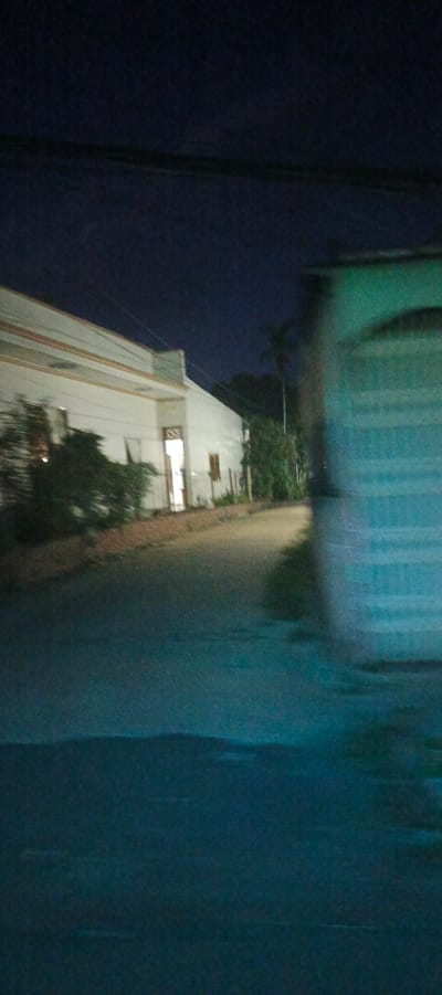 This image depicts a blurry, low-light scene at night, likely on a street in Xuân Long, Vietnam. The dominant features are a long, white building with a light emanating from an open doorway on the left, and a blurred, teal-colored structure on the right. Trees and foliage are visible along the side of the white building. The road in the foreground is dark and appears wet, reflecting some light. A power line stretches across the top of the frame. The overall impression is one of motion, possibly captured from a moving vehicle.
