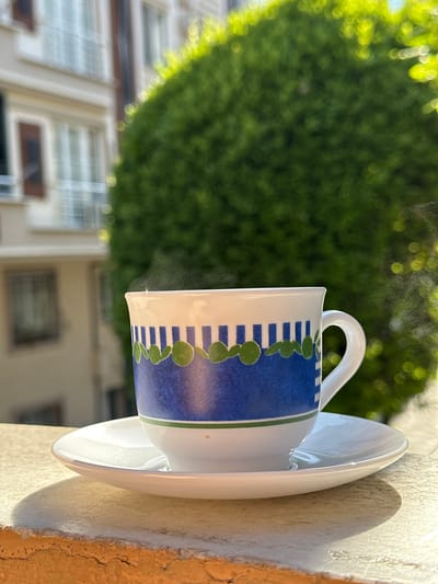 A close-up shot shows a white coffee cup with a blue and green patterned band, sitting on a matching saucer. Steam rises from the coffee. The cup and saucer are placed on a textured, light-colored ledge. In the background, a blurred building with windows and a large, out-of-focus green bush are visible, suggesting an outdoor setting, possibly in Bahçelievler, Turkey. Sunlight casts a shadow from the cup and saucer onto the ledge.