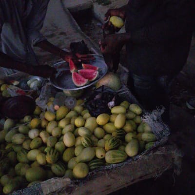 In Wuro Allahamdu, Nigeria, a vendor, partially obscured by shadow, is seen preparing fruit. Sliced watermelon rests on a metal plate, while other fruits, possibly melons or gourds, are piled in a sack. The ground appears to be dirt. The scene suggests an outdoor market or roadside stall, illuminated by dim, possibly artificial, light.