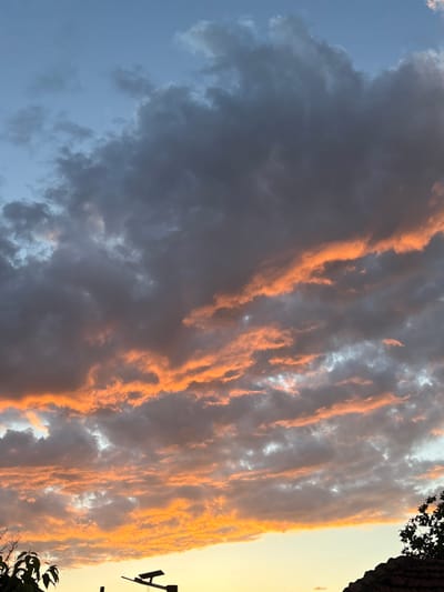 The image captures a dramatic sky scene, likely at sunset or sunrise, over Pchelnik, Bulgaria. The central focus is a vast expanse of clouds stretching across the frame, characterized by a striking contrast between dark, voluminous forms and intensely illuminated golden-orange segments.
On the left side of the image, the sky is a soft, clear blue, gradually transitioning into darker, heavier grey and blue-grey clouds that dominate the mid-ground. These clouds appear dense, suggesting a recent storm or heavy weather conditions. Interspersed and running vertically through the darker cloud mass are elongated, fiery streaks and patches of clouds, brilliant with warm hues of orange and gold. This vibrant illumination indicates the low angle of the sun, casting a radiant glow on the cloud edges and upper surfaces.
Towards the far right, the sky brightens significantly to a clear, luminous golden-yellow, hinting at the sun's position just outside the frame. In the bottom and top right corners, dark silhouettes of tree branches with foliage are visible against this bright backdrop, along with what appears to be a portion of a dark building or roofline.
There are no people or discernible activities depicted. The scene emphasizes the natural beauty and power of the sky, creating a visually compelling and almost painterly effect with its blend of intense colors and dramatic cloud formations. No text is visible in the image.
