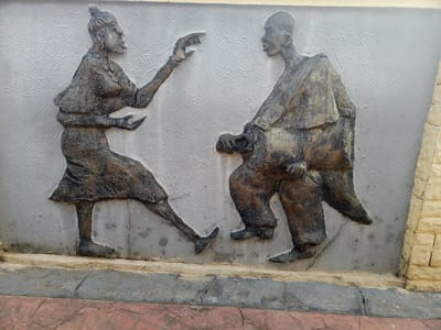 This media file features a relief sculpture on a light gray, textured wall, depicting two human figures in dark, earthy tones.
On the left, a female figure is shown in profile, facing right. She has her hair tied up and wears a dress or skirt. Her left arm is extended with an open hand, suggesting a gesture of speaking or emphasis, while her right hand is held lower, near her waist. She appears to be in motion or actively communicating.
On the right, a male figure is also depicted in profile, facing left, looking towards the woman. He appears to be seated or in a stooped posture, dressed in loose robes or traditional attire, and has a bald or shaven head. He seems to be listening attentively to the woman.
The interaction between the two figures suggests a narrative, possibly a conversation, a storytelling session, or a depiction of a traditional dance or cultural exchange. The sculpture itself has a rough, textured finish, giving it an aged or weathered appearance. Below the wall, there is a paved ground with sections of reddish and gray tiles. A portion of a light yellow brick wall is visible on the far right. The scene is outdoors and illuminated by natural daylight. No visible text is present in the image. The style and figures are indicative of the cultural context of Osogbo, Nigeria.