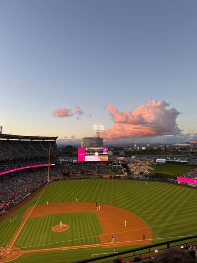 The image captures an elevated view of a baseball game in progress at Angel Stadium in Anaheim, United States, during sunset. The sky displays a serene gradient from light blue overhead to warm pink and orange hues on scattered cumulus clouds, illuminated by the setting sun. Stadium lights are already on, casting a glow over the scene.

The central subject is the baseball field, with its meticulously striped green grass outfield and infield, contrasting with the reddish-brown dirt of the baselines and pitcher's mound. Several baseball players are visible on the field, including a pitcher on the mound, a batter at home plate, and fielders positioned around the bases, indicating an active game. The multi-tiered stadium seating is filled with numerous spectators, particularly on the first base side (left of the image), suggesting a well-attended event.

Notable stadium features include a large video scoreboard in the center-left outfield displaying a baseball graphic and the "T-Mobile" logo. To its right, a distinctive rock formation resembling mountains, a signature element of Angel Stadium, is visible behind the outfield wall. Various advertisements line the outfield fence and are visible on screens and buildings. Text visible includes "Angels" on a building in the background, "T-Mobile," "Modelo," "Yakult," "Coca-Cola," "Toyota," "MemorialCare," "National," and "Pechanga Resort Casino" on a smaller screen to the right. The overall impression is a vibrant evening at a professional baseball game under a striking sunset.
