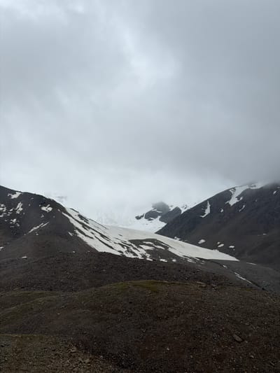 An overcast, grey sky looms over a rugged mountain landscape. The foreground is a steep, rocky slope, transitioning into a higher mountain range with patches of snow scattered across its dark, stony surface. Further in the distance, a vast, snow-covered mountain peak emerges, partially obscured by mist and clouds. The scene is devoid of any human presence or discernible structures, suggesting a remote, natural environment.
