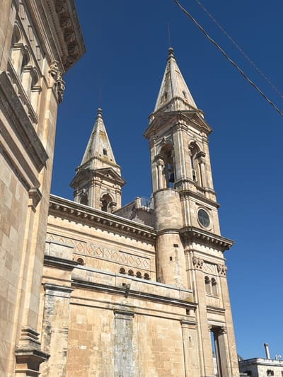 This image captures a section of a large, historic stone building, likely a church or cathedral, under a clear blue sky. The architecture is characterized by intricate stone carvings, arched windows, and decorative friezes. Two prominent bell towers with pointed spires rise from the structure, one taller and more ornate than the other. The stonework appears weathered, showing patches of discoloration and moss. In the foreground, to the left, a portion of another stone building with similar architectural elements is visible, creating a layered perspective. Electrical wires are faintly visible in the upper right portion of the frame, crossing the sky. The image is taken from a low angle, emphasizing the height and grandeur of the buildings. The location is identified as Alberobello, Italy.