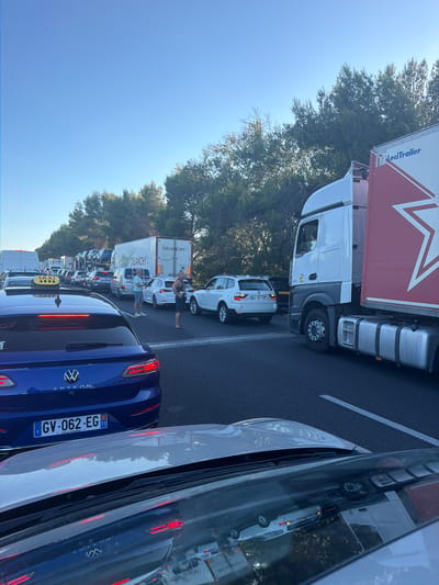 The image depicts a significant traffic jam on a highway in Villeneuve-lès-Béziers, France. The scene is dominated by a line of stationary vehicles, including cars, vans, and trucks. In the foreground, the rear of a blue Volkswagen Arteon is visible, with its license plate reading "GV-062-EG". To the right of the Arteon, a white BMW X5 is stopped. Further down the highway, a large white semi-trailer truck with a red stripe and a logo that appears to read "LeciTrailer" is stationary. Next to it, a white box truck with "DELANCHY" branding is also stopped. Two men are standing on the side of the road, one in a light blue shirt and the other in a dark tank top, seemingly observing the traffic situation. The sky is clear and blue, and trees line the side of the highway. The road surface is asphalt with white lane markings. The overall impression is one of a standstill due to a traffic obstruction.