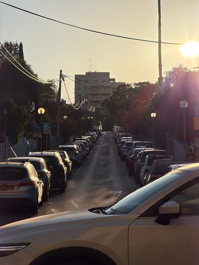 The image captures a sun-drenched street scene in Kfar Saba, Israel, at either sunrise or sunset, indicated by the low, brilliant sun creating prominent lens flare and long shadows. The street extends from the foreground into the distance, lined with numerous cars parked bumper-to-bumper on its left side. A large white car occupies the right foreground, with part of its side and mirror visible.
The golden light illuminates the asphalt road, creating reflections, and highlights the tops of the parked vehicles. Beyond the cars, on the left, are trees and foliage, some bathed in the warm light. In the background, a multi-story, light-colored building with visible windows stands amongst the greenery. Overhead, against a clear, bright sky, utility poles and a network of power lines traverse the frame.
Notable details include a blue sign in the middle ground featuring a white wheelchair symbol, indicating accessible parking. A smaller, less distinct sign is positioned below it. The license plate of a silver car further down the street is partially visible, showing the numbers "23-75." The scene portrays a tranquil urban or suburban setting, largely static with no explicit human activity, dominated by the warm, directional light of the low sun.
