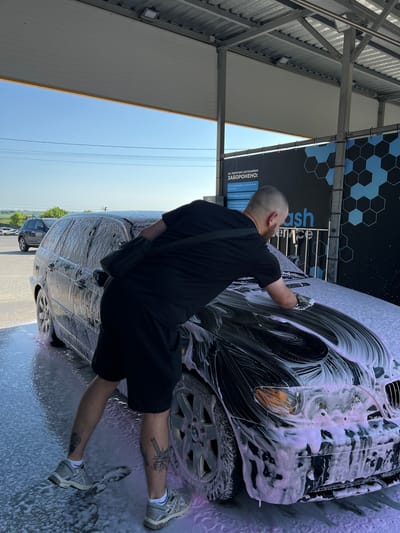 A man is washing a dark-colored station wagon at a self-service car wash in Velyki Hai, Ukraine. The man is wearing a black t-shirt and black shorts, with tattoos on his legs. He is using a sponge to apply foam to the car. The car is covered in pink and white soap suds. The car wash is an outdoor facility with a metal roof and posts. A sign in Ukrainian is visible in the background with the text "НА ТЕРИТОРІЇ АВТОМИЙКИ ЗАБОРОНЕНО:" which translates to "ON THE TERRITORY OF THE CAR WASH, IT IS FORBIDDEN:". The sky is clear and blue. Another car is parked in the background.
