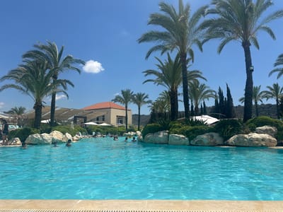 The image depicts a resort swimming pool scene in Qraiyeh, Lebanon. A large, turquoise swimming pool occupies the foreground, with its edge visible at the bottom of the frame. Several people are in the pool, some partially submerged and others swimming. The background features lush greenery, large rocks, and palm trees, characteristic of a Mediterranean resort. A building with an orange roof is visible behind the greenery, and white umbrellas and lounge chairs are scattered around the pool area, suggesting a recreational setting. The sky is a clear blue with a few scattered clouds.