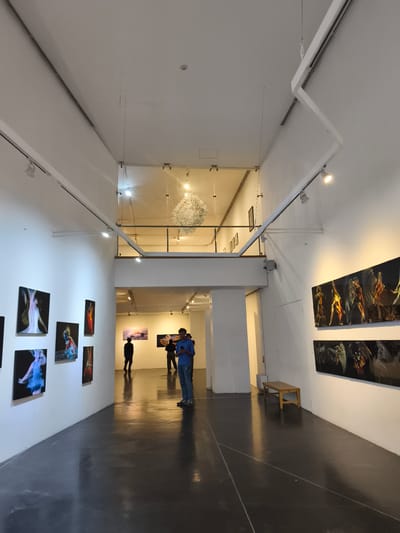The image depicts the interior of an art gallery in Querétaro, Mexico. The space is characterized by white walls, a polished dark gray floor, and high ceilings with exposed ductwork and track lighting. A mezzanine level overlooks the main gallery space, with a decorative white spherical chandelier suspended from the upper ceiling. Various pieces of artwork are displayed on both walls, including photographic prints and paintings. Several people are present in the scene. One man stands in the foreground, facing away from the camera, looking at his phone. Two other individuals are further down the gallery in another gallery space, possibly viewing the art as well. A wooden bench sits against the right wall of the gallery.