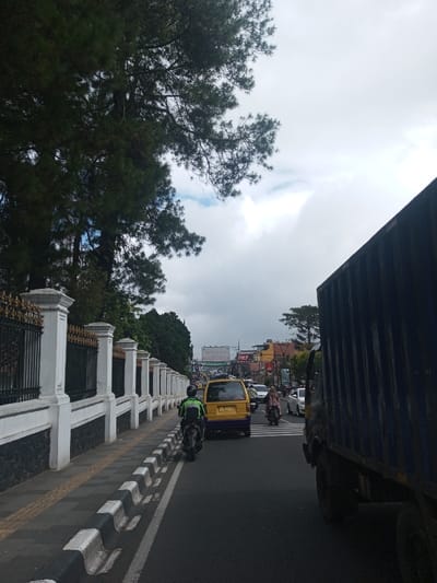 The scene, located in Thotagamuwa, Sri Lanka, depicts a busy urban street under an overcast sky. In the immediate foreground on the right, a dark blue commercial truck's cargo box and rear wheel are partially visible, indicating its presence in the rightmost lane or shoulder. To the left, a pedestrian sidewalk runs alongside the roadway, bordered by a decorative white concrete fence featuring regularly spaced pillars and ornate black wrought iron panels with gold embellishments. Tall evergreen trees with needle-like leaves rise prominently in the upper left portion of the frame.
The asphalt road features a black-and-white striped concrete barrier or curb segmenting the shoulder from the main traffic lanes. A motorcyclist wearing a green jacket is positioned just past this barrier on a dark grey motorcycle. Further down the road in the midground, a yellow compact van with a visible rear license plate is present in a traffic lane. To its right, another motorcyclist, partially obscured and appearing to wear a head covering, is situated on what appears to be a pedestrian crossing marked by diagonal white lines on the road. Multiple additional vehicles, including cars and other light vehicles, are visible further in the distance, indicating active traffic. The background shows a collection of buildings, utility poles, streetlights, and billboards, suggesting a developed commercial or residential area.