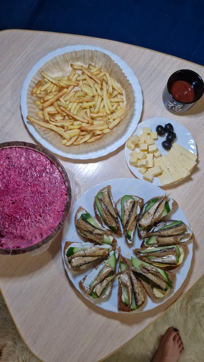 A high-angle shot shows a light-colored wooden table laden with food. There is a small black bowl containing a red sauce in the upper left. Adjacent to it is a white plate featuring slices and cubes of cheese, accompanied by a small pile of black olives. Below these, a white plate filled with french fries sits next to a large glass bowl filled with pink salad. To the right, there is another white plate containing open-faced sandwiches topped with cucumber and fish. A human foot is partially visible in the upper right of the frame. The location is presumed to be Otchor, Russia, based on provided metadata.