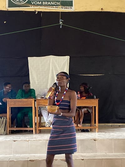 The image depicts an indoor scene with several people, likely in Nigeria given the location context. In the foreground, a young person wearing a striped skirt and a beaded necklace is holding a microphone and what appears to be a small instrument or prop. They are looking slightly to the side. Behind this individual, a white cloth is draped, and further back, another person is visible sitting, partially obscured by a wooden structure. To the left of the foreground subject, a sign with text is partially visible. The text reads "VOM BRANCH" and some numbers. The setting appears to be a stage or performance area, with a dark backdrop. The lighting suggests an indoor event. The mood seems to be attentive, possibly during a performance or presentation. The overall scene is dimly lit, but the focus is on the individuals and the activity taking place.