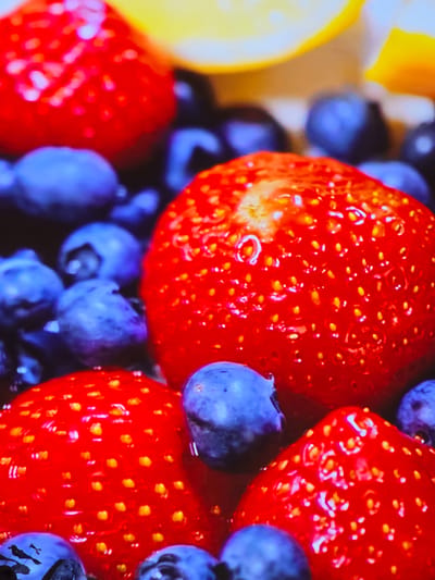 The image is a close-up, vibrant, and slightly out-of-focus shot of a bowl of mixed berries, primarily strawberries and blueberries. The lighting is bright, highlighting the glossy surfaces of the fruits, with a bright yellow element, likely a slice of lemon or orange, visible in the upper portion of the frame. The focus is sharpest on the central strawberry and surrounding blueberries. There are no people, actions, or environmental details of Mawindi, Tanzania, depicted in this image.