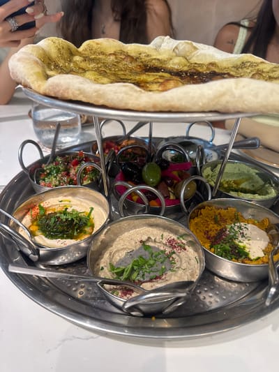 An overhead shot shows a platter of Middle Eastern appetizers at a table in Aventura, Florida. The platter is made of stainless steel and features a raised section in the center, holding a flatbread topped with herbs and oil. Arranged around the central section are several small metal bowls filled with various dips and sides. These include hummus with a drizzle of green sauce and red pepper flakes, a creamy dip topped with herbs and red spice, a yellow dip with a swirl of white sauce and a sprinkle of herbs, a green dip, a salad with chopped tomatoes and parsley, and a selection of olives and pickled vegetables. The platter is placed on a white marble-patterned table. In the background, two women are partially visible, with one in the foreground holding a dark object, possibly a phone. A glass of water is visible to the left of the platter.