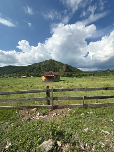 The image depicts a rural landscape with a traditional wooden house and several yurts in a grassy field. A rustic wooden fence runs across the foreground. In the background, a verdant, tree-covered mountain rises under a dramatic sky with large, white clouds. The scene is set in the village of Kazanovka, Russia, in a natural environment.
