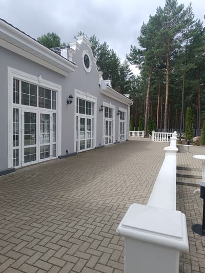 The image shows a light gray building with large white-framed windows and doors, situated in an outdoor paved area. Tall pine trees are visible behind the building. The paving is made of interlocking grey bricks laid in a herringbone pattern. A white railing with decorative balusters runs along the paved area, separating it from a slightly elevated section with steps and white tables. The sky is overcast. The building appears to be a restaurant or event venue, possibly located in a wooded area on the outskirts of a settlement. The scene suggests a quiet, possibly off-season day. No people are visible in the image.