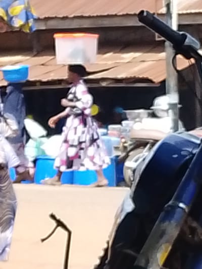 The image depicts a street scene in Otulu, Nigeria, likely during daylight hours with a bright, possibly sunny, atmosphere. In the foreground, a portion of a dark blue motorcycle or bicycle is visible, with its handlebars and part of the frame taking up the right side of the frame. A thin, dark, U-shaped metal object, possibly part of a stand or railing, is also in the lower left corner.
The middle ground features a woman walking. She is wearing a patterned dress with black, white, and pink geometric designs and is balancing a large, clear plastic container with an orange lid on her head. She is surrounded by various objects, including blue barrels or buckets, and what appear to be bundles of goods wrapped in plastic or fabric. Behind her, another person, possibly a child, is also seen with a blue bowl balanced on their head, and further back, another figure in a dark blue garment. The background consists of stalls or shops with corrugated metal roofs.
The scene suggests a busy market or roadside trading environment where goods are transported and sold. The overall impression is one of daily commerce and activity. There is no visible text in the image.