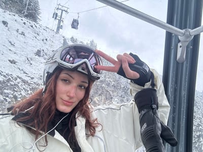 The image depicts a young adult female inside a ski lift gondola, located in a ski resort within the commune of Les Allues, France. The female, positioned centrally, has long, wavy reddish-brown hair and a slight smile. She wears a light-colored ski helmet with white-framed ski goggles pushed up onto the helmet. Her attire includes an off-white ski jacket over a black high-necked base layer. Her right hand, wearing a black liner glove or partial glove, forms a peace sign with its bare index and middle fingers. Her left hand, fully covered by a black ski glove featuring a visible "REUSCH" logo and wrist strap, rests by her side. The immediate right background shows the interior metal framework of the gondola cabin. Through the transparent window, a snow-covered mountainous landscape is visible under an overcast sky, featuring scattered pine trees. In the mid-ground, a prominent ski lift tower with multiple rollers is observed, along with two other black-colored enclosed gondola cabins suspended from the ski lift cable system.