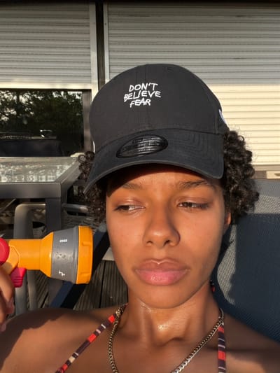 The image shows a close-up of a young woman wearing a black baseball cap that says "DON'T BELIEVE FEAR". She is holding an orange and grey garden hose nozzle. Her face is partially illuminated by the golden hour sun. The background suggests an outdoor setting, possibly a backyard, with a roller shutter door and some furniture visible. There is a slight sheen of sweat on her neck and upper chest, indicating warm weather. The scene is likely in the city of Champs-sur-Marne, France, given the prompt.