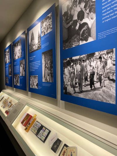 The image shows a museum exhibit in Banguntapan, Indonesia. It is a white-walled room with several framed black and white photographs and several display cases containing various objects. The photos are all in a similar style with a blue border.  The photos depict historical scenes, including a group of people in traditional clothing, a procession of people in a town, a group of people standing in front of a building, and a man in a suit and tie walking on a path. There is a caption below each picture, though they are not visible in the image.  There are multiple glass display cases with various artifacts and objects on white-covered tables running along the wall.  Some of the artifacts include several cards of playing cards, two sets of playing cards, a card game, a small wooden box with red contents, and a red box containing playing cards. The display cases have a glass top, and the room appears to be well-lit.