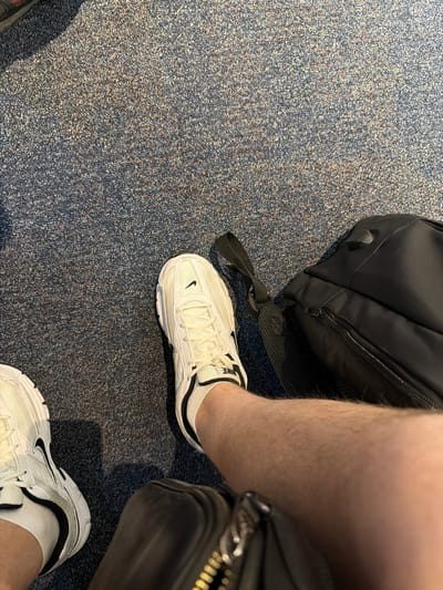 The image shows a person's legs wearing white Nike sneakers and white socks. The person is sitting on a blue carpeted floor, possibly in an airport. Next to the person is a black backpack. The location is the Aeropuerto Internacional Ezeiza, Argentina.