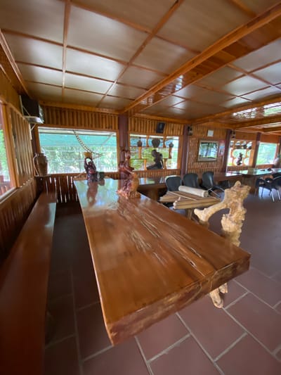 The image depicts the interior of a restaurant with a rustic, natural theme, likely located in Thôn 2, Vietnam. The dominant material is wood, used for the ceiling, walls, and furniture. The ceiling is a grid of wooden beams with panels in between, giving it a segmented appearance. The walls are constructed from vertical bamboo poles.
A prominent feature in the foreground is a large, polished wooden table with an irregular, natural edge, supported by a unique wooden leg that resembles a gnarled tree root. Adjacent to this table is a long, wooden bench seat. On the table, there are several decorative figurines, including one of a person holding a bowl above their head and another figure that appears to be a statue of a person sitting. A large, dark brown ceramic vase is visible on a ledge to the left of the table.
In the background, more wooden tables and chairs are visible. These tables are also made of polished wood, and the chairs are black. Large windows with a green, mesh-like covering look out onto an outdoor, leafy environment, suggesting a garden or natural setting. The floor is tiled with reddish-brown square tiles. Several decorative items and lamps are mounted on the walls. The overall impression is of an open-air or semi-open-air dining space designed to blend with nature.