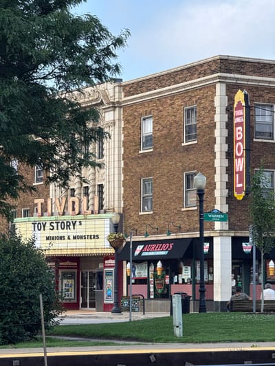 The image captures a street scene in Downers Grove, Illinois, featuring the historic Tivoli Theatre and adjacent businesses. The Tivoli Theatre marquee displays "TIVOLI" in neon lights above "TOY STORY 5" and "MINIONS & MONSTERS". To the right of the theatre, a vertical neon sign reads "TIVOLI EXPRESS BOWL". Below the theatre marquee, the entrance to Classic Cinemas is visible, with movie posters displayed. Next to the theatre, Aurelio's The Family Pizzeria has its awnings and signage. A street lamp with a "WARREN AVENUE" sign stands between the theatre and the pizzeria. Two individuals are seated on a park bench in front of Aurelio's, facing away from the camera. The scene is set against a backdrop of a brick building with white architectural details and a clear sky overhead. A lush green tree partially obscures the left side of the theatre.