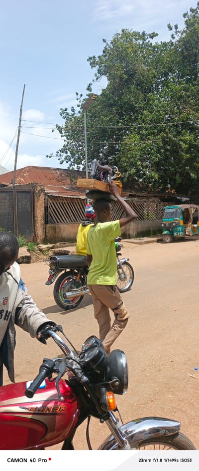 In Barikin Gidar Kanat, Nigeria, a young person in a light green t-shirt walks along a dirt road, balancing a sewing machine on their head. The sewing machine, mounted on a wooden base, is secured with padding. Behind them, another person in a yellow shirt sits on the back of a motorcycle. Further down the road, a turquoise and green auto-rickshaw, with the license plate "PT 16493," is parked. The background features a building with a corrugated iron roof and a fence made of wooden slats. Lush green trees line the street, and the sky is partly cloudy. In the foreground, the handlebars and front of a red motorcycle are visible.