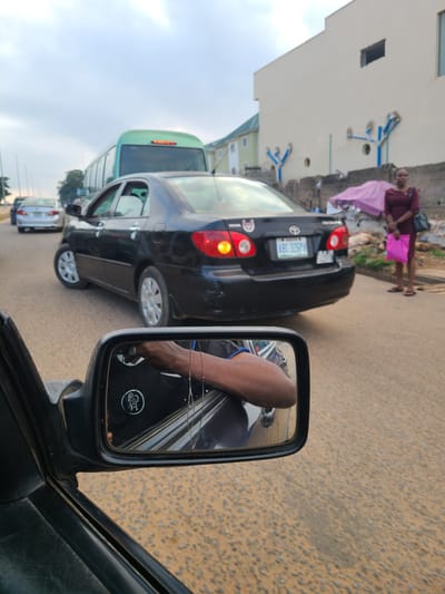 In Karu, Nigeria, traffic moves slowly under an overcast sky. The viewpoint is from inside a vehicle, looking out at the road and through a side mirror. A black sedan is the most prominent vehicle, with its brake lights illuminated. Beyond it, a light-colored sedan is visible, followed by a green bus. To the right of the road, a woman in a burgundy dress stands holding a pink bag. The side mirror reflects the arm and hand of the driver, a black shirt with a white logo, and the interior of the car. The road surface appears to be asphalt with visible aggregate. Buildings are visible in the background, with one being a light-colored, multi-story structure.