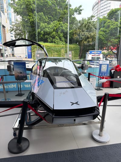 A white and black eVTOL (electric vertical takeoff and landing) vehicle is displayed on a platform with wheels, enclosed by stanchions and red velvet ropes, inside a building with large glass windows. The vehicle has a futuristic design with a glass canopy and visible rotors. To the left, a poster in Chinese depicts a similar vehicle over a cityscape. Behind the vehicle, through the glass, a street scene is visible with trees, buildings, and a police vehicle. A QR code sign with Chinese text is also present. The overall environment suggests an indoor exhibition or showroom. The scene is located in Futian District, China.