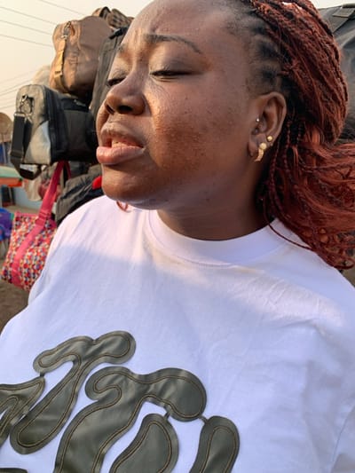 A close-up depicts a woman with reddish-brown braided hair styled in a bun. Her skin tone is dark, and she has multiple ear piercings adorned with gold jewelry. Her eyes are closed. She's wearing a white t-shirt featuring an abstract pattern or design in a muted green or olive tone. A dark-colored bag or backpack rests against her, and a brightly colored woven bag is visible in the background.