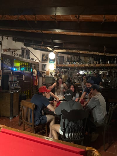 The image captures a dimly lit, interior shot of what appears to be a bar or restaurant in Lequeitio, Mexico. The establishment features a rustic decor with exposed brick, dark wooden beams, and beer coasters. Patrons are sitting and standing around tables, possibly engaging in conversations and socializing. A pool table with a red surface is also visible. The walls are adorned with various framed images, hats, and decorative elements. Slot machines are against a wall.