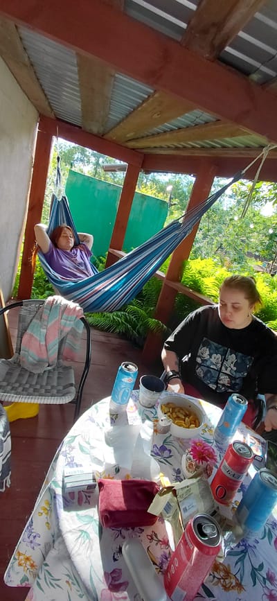 The image depicts a shaded outdoor patio with two individuals. In the background, a woman with red hair lies in a striped hammock, resting with her hands behind her head. In the foreground, a woman with brown hair, wearing a black t-shirt with cartoon characters, sits at a table covered with a floral tablecloth. Various items are spread on the table, including several cans of beverages, a bowl of what appears to be food, a pack of "HEETS," and a folded piece of fabric. The patio is constructed with wooden beams and a corrugated metal roof. Lush green foliage is visible in the background, suggesting a natural, possibly rural or semi-rural, setting. The lighting indicates it is daytime. There is no visible indication of the location being Tashkazgan, Russia.