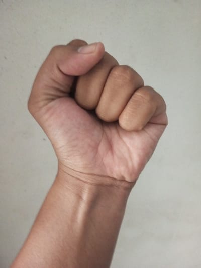 A closed fist, oriented vertically, is shown against an off-white background. The hand is tan, transitioning to a darker shade at the knuckles. Veins are subtly visible on the wrist and forearm. Skin wrinkles are present on the back of the hand and around the wrist. The thumb is positioned over the closed fingers. The overall impression is one of tension or potential strength.