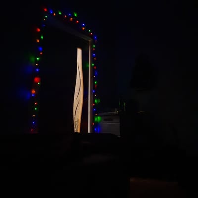 An interior scene in Kurganinsk, Russia, depicts a dimly lit room with a partially open doorway serving as the primary light source and focal point. The doorframe is adorned with a string of multicolored LED lights, displaying red, green, and blue illumination in an alternating pattern. The doorway itself reveals a sliver of a brightly illuminated space beyond, with a light-colored, possibly wooden, door panel featuring a curved decorative element. To the right, within the room beyond the door, a light-colored object, potentially a piece of furniture, is faintly visible, along with diffuse blue and red indicator lights from an unseen electronic device. The foreground room from which the image is taken is predominantly dark, with no discernible individuals or specific objects. No actions or interactions are observed. The environment suggests an indoor, possibly domestic, setting under low-light conditions.