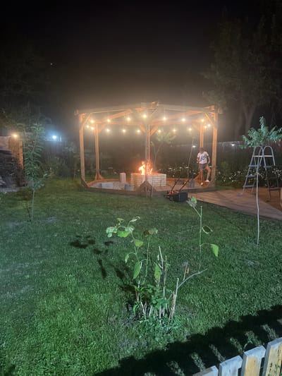 The image depicts a backyard scene at night in Kok Jar, Kyrgyzstan. A young person, wearing a white t-shirt with blue lettering and denim shorts, stands near a wooden gazebo. The gazebo is illuminated by a string of lights and features an active fire pit with flames. A red and black lawnmower is positioned on a wooden deck adjacent to the gazebo. The ground is covered in green grass, and there are various plants and trees surrounding the area. A wooden fence runs along the bottom edge of the frame. The sky is dark, with some faint lights visible in the distance.