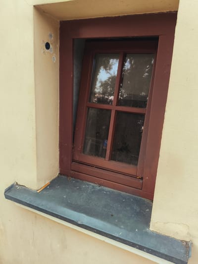 The image shows a close-up view of an old, slightly dilapidated window set into a light yellow stucco wall. The window has a dark reddish-brown wooden frame with a traditional cross-pane design. One of the window panes is slightly ajar, revealing reflections of trees and sky, suggesting it is daytime and the weather is clear. The window sits above a dark grey metal or stone windowsill, which appears weathered and has some debris on it. To the left of the window frame, on the stucco wall, there is a small, dark, circular object, possibly a security camera or a light fixture, surrounded by chipped paint and plaster. The overall impression is one of age and a rural or historical setting, consistent with a location in Nowy Dwór Mazowiecki, Poland. No people or specific events are visible. The scene evokes a sense of quietness and a focus on architectural details and the passage of time.