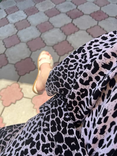 The image displays a close-up view of a person's foot wearing a light-colored sandal, positioned on a patterned tiled surface. The tiles are arranged in a checkerboard pattern with alternating light pink and light gray cloud-shaped segments. The person is also wearing a garment with a black and white leopard print pattern, which is prominently featured in the foreground, partially obscuring the foot and tiles. The lighting suggests an outdoor setting, with a shadow cast on the tiles to the left of the foot. The location is identified as Qoyankus, Kazakhstan.