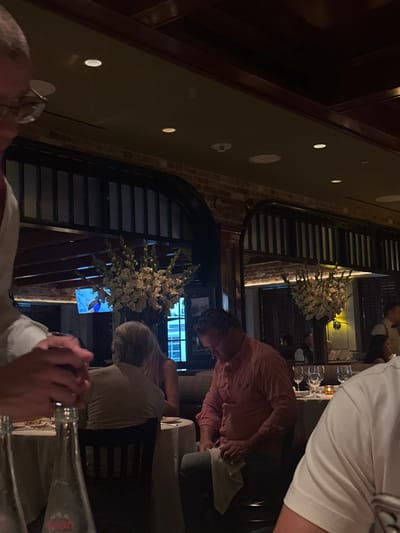 The image depicts an indoor restaurant environment in University Park, United States. The scene is characterized by dim, warm lighting from recessed ceiling fixtures and table candles. The interior features exposed brickwork, dark wooden archways, and paneled elements. Large floral arrangements, primarily white gladiolus-like flowers, are prominently displayed throughout the dining area.
In the foreground, the upper torso, head, and hands of a person wearing a white shirt and glasses are visible on the left, holding a dark glass or bottle. Two clear glass bottles, one labeled "Evian," rest on a white-clothed table. On the far right foreground, a partial view of a person's shoulder and arm in a white shirt is visible.
In the mid-ground, a male subject wearing a light orange or salmon-colored long-sleeved collared shirt is seated at a table, looking down and actively wiping his hands with a white cloth. To his left, another individual with gray hair, viewed from the back, is seated at a table alongside a person with blonde hair, also seated. Multiple white-clothed dining tables are arranged, each set with multiple wine glasses, and some feature lit candles. In the background, a television screen displays a blue and white image, likely a sporting event, and additional indistinct figures of diners and restaurant staff are visible.